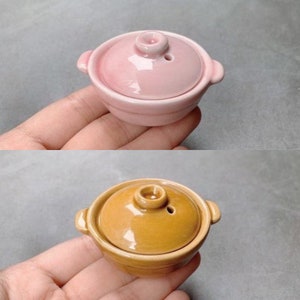 May include: Two small ceramic pots with lids. The pot on the top is pink and the pot on the bottom is brown.