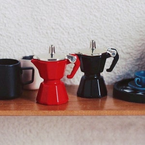Pode incluir: Duas pequenas cafeteiras de espresso, uma vermelha e outra preta, com tampas e alças prateadas. As cafeteiras estão sobre uma superfície de madeira.
