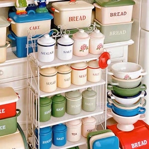 以下が含まれることがあります： 白い金属製の棚ユニットで、複数の棚にミニチュアのキッチン収納容器が置かれています。容器には「砂糖」、「塩」、「お茶」、「コーヒー」などの言葉が書かれています。棚には、ミニチュアのパン箱、ケーキ容器、ボウルも置かれています。