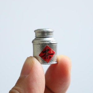 Puede incluir: Un pequeño recipiente de metal plateado con una etiqueta roja y negra que dice "茶" en caracteres chinos. El recipiente está siendo sostenido en la mano de una persona.