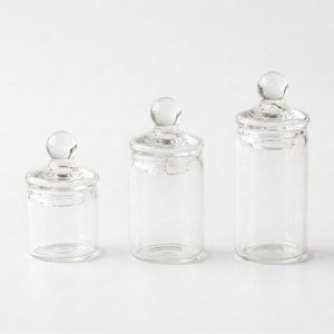 Puede incluir: Tres botes de farmacia de vidrio transparente con tapas redondeadas. Los botes varían en altura, con un bote pequeño, mediano y alto. Cada bote tiene un pomo redondo en la parte superior de la tapa.