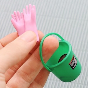 May include: A miniature green bucket with a handle and a small label, next to a pair of pink gloves. The items are small and appear to be toys or part of a playset.