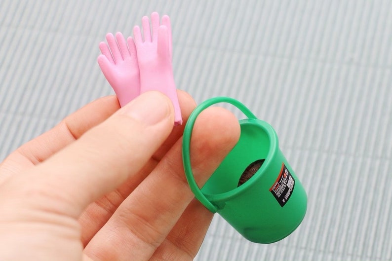 May include: A miniature green bucket with a handle and a small label, next to a pair of pink gloves. The items are small and appear to be toys or part of a playset.