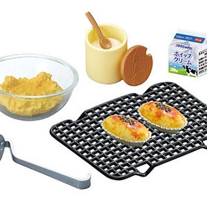 Op de afbeelding: Een miniatuurkeuken speelset met verschillende accessoires, waaronder een kom met gele deeg, een klein bakje met room, een houten deksel, een kleine metalen stamper en een zwart metalen bakrooster met twee kleine gebakken producten.