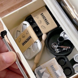 May include: A collection of miniature baking tools and accessories, including small loaf pans, a rolling pin, measuring spoons, and a muffin tin. The items are packaged in a white drawer with the word "BAKEWARE" printed on the packaging.