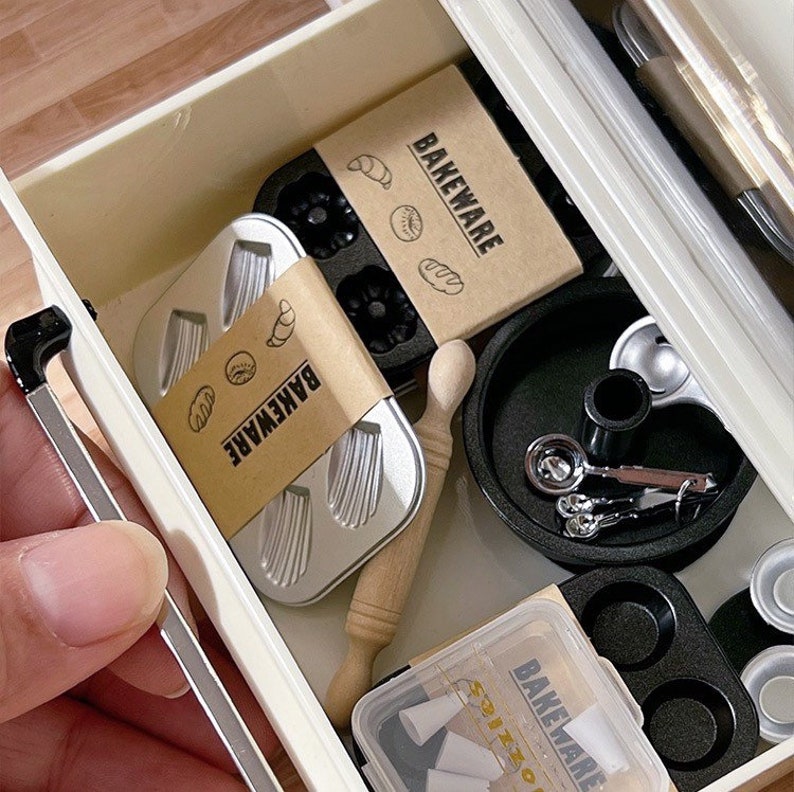 May include: A collection of miniature baking tools and accessories, including small loaf pans, a rolling pin, measuring spoons, and a muffin tin. The items are packaged in a white drawer with the word "BAKEWARE" printed on the packaging.