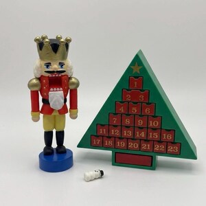 Könnte beinhalten: Eine hölzerne Nussknacker-Figur mit goldener Krone, roter Jacke und gelber Hose auf einem blauen Sockel, neben einem grünen, dreieckigen Adventskalender mit dreiundzwanzig nummerierten roten Schubladen und einer kleinen weißen Schneemann-Figur.