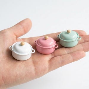 May include: Three miniature, colorful pots with lids. One is white, one is pink, and one is mint green. Each pot has small handles and a wooden knob on the lid. The pots are held in a person's hand.