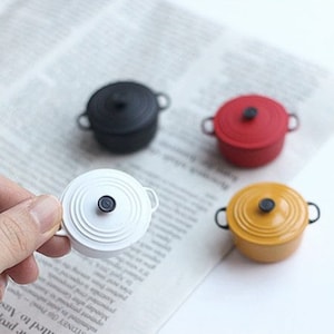 May include: Miniature colorful cast iron pots in black, red, and yellow, with a white pot held by a hand. Each pot has a lid and small handles. The pots are on a newspaper background.