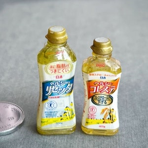 Può includere: Due bottiglie di olio da cucina in plastica trasparente. La bottiglia a sinistra è etichettata "Olio sano" e la bottiglia a destra è etichettata "Colesterolo sano". Entrambe le bottiglie sono gialle e hanno un tappo dorato.