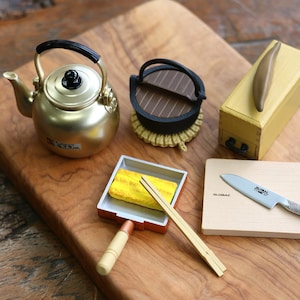 Könnte beinhalten: Ein Miniatur-Küchenset mit einem goldenen Wasserkocher, einem kleinen schwarzen Grill, einem Holzbrett mit einem Messer, einer kleinen Bratpfanne mit einem Ei und Essstäbchen sowie einer Holzkiste mit einem braunen Gegenstand.