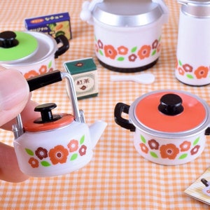 Puede incluir: Un juego de utensilios de cocina en miniatura con un patrón floral. El juego incluye una tetera, una olla arrocera, una olla con tapa y un termo. Todos los artículos son blancos con diseños florales naranjas y marrones.