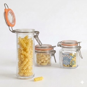 Op de afbeelding: Drie heldere glazen Kilner potten met oranje deksels en metalen sluitingen, gevuld met pasta. De grootste pot is open en onthult fusilli pasta. De andere potten bevatten verschillende pastavormen. De potten staan op een wit oppervlak.