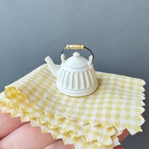 Op de afbeelding: Witte keramische theepot met een zwart handvat en gouden accenten op een geel en wit geruit tafelkleed.