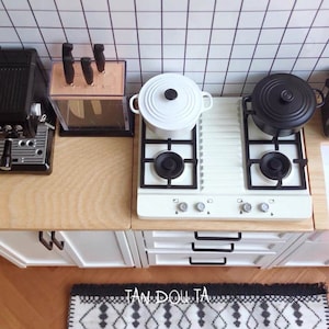 May include: Miniature kitchen scene with a white stove, black pots, and a coffee maker. A knife block and a patterned rug are also visible. The kitchen has a white tile backsplash and wooden countertops.