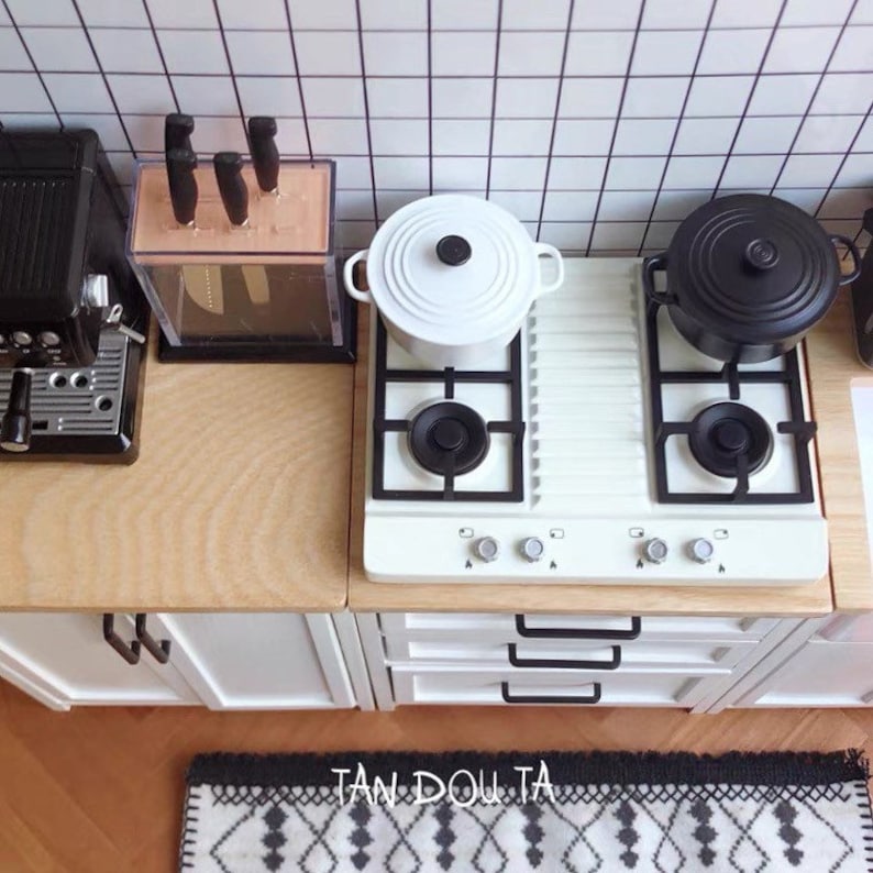 May include: Miniature kitchen scene with a white stove, black pots, and a coffee maker. A knife block and a patterned rug are also visible. The kitchen has a white tile backsplash and wooden countertops.