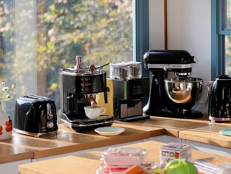 Peut inclure: Une collection d'appareils de cuisine noirs, dont un grille-pain, une machine &agrave; expresso, une cafeti&egrave;re, un batteur sur socle et une bouilloire, est dispos&eacute;e sur un comptoir en bois. Une tasse blanche est sous la machine &agrave; expresso.