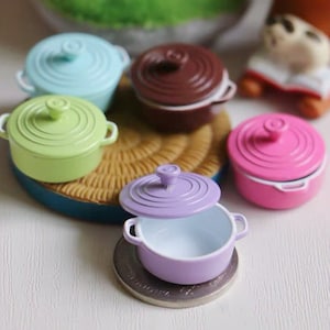 May include: A collection of miniature, colorful ceramic pots with lids. The pots are in shades of blue, green, brown, pink, and purple. One purple pot has its lid slightly ajar, revealing a white interior. The pots are arranged on a white surface.