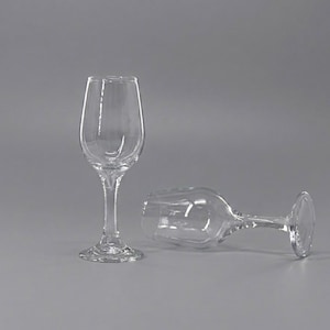 May include: Two clear, stemmed wine glasses. One glass stands upright, while the other lies on its side. The glasses have a classic design with a rounded bowl and a slender stem. The base of the upright glass has a decorative pattern.