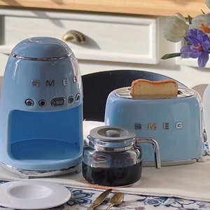 Peut inclure: Ensemble d'appareils de cuisine SMEG bleu clair comprenant une cafeti&egrave;re, un grille-pain avec une tranche de pain grill&eacute; et une carafe en verre. Une assiette blanche et des couverts sont pos&eacute;s sur la table.