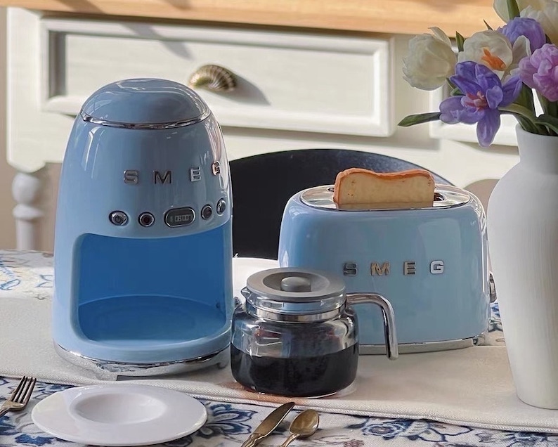 Peut inclure: Ensemble d'appareils de cuisine SMEG bleu clair comprenant une cafeti&egrave;re, un grille-pain avec une tranche de pain grill&eacute; et une carafe en verre. Une assiette blanche et des couverts sont pos&eacute;s sur la table.
