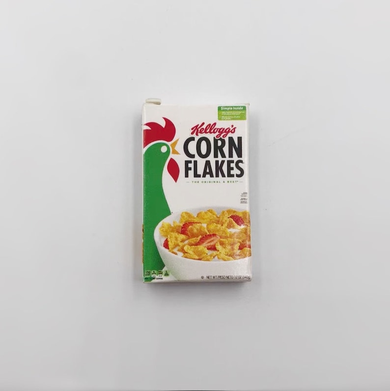 May include: A small box of Kellogg's Corn Flakes cereal. The box features a green rooster graphic and the words "Kellogg's CORN FLAKES" in red and black. A bowl of cereal with strawberries is pictured on the front. The box is 12 oz.