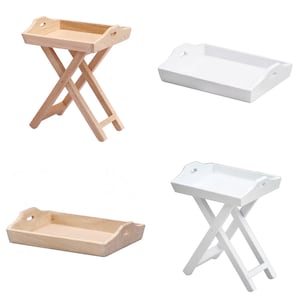 Könnte beinhalten: Zwei Holz-Klapptische mit Tablett, einer in Naturholz und einer in Weiß. Beide Tische haben eine rechteckige Tablettplatte mit abgerundeten Kanten und zwei Griffen an den Seiten. Die Tische sind zusammengeklappt und stehen auf einem weißen Hintergrund.
