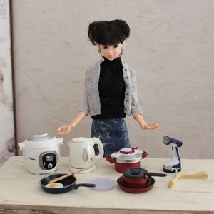 Op de afbeelding: Een pop met een grijze trui en blauwe jeans staat voor een tafel met een verscheidenheid aan miniatuurkeukengerei, waaronder een witte elektrische waterkoker, een witte rijstkoker, een rode snelkookpan, een koekenpan en een blauw-wit strijkijzer.
