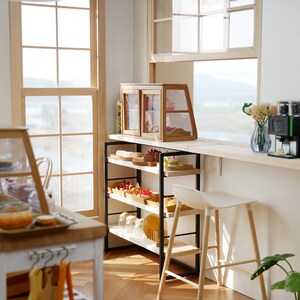 May include: A miniature bakery shop interior with a white counter, wooden shelves, and glass display cases filled with pastries. A coffee maker and toaster are on the counter, and a white stool sits next to the counter.