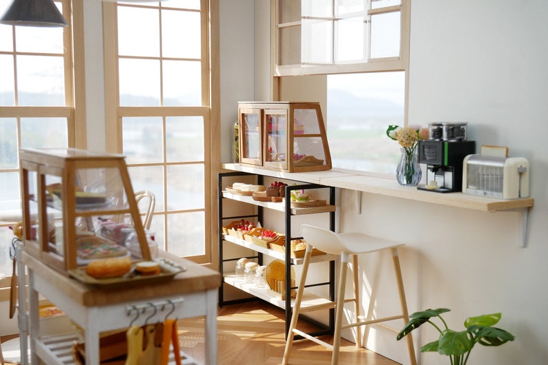 May include: A miniature bakery shop interior with a white counter, wooden shelves, and glass display cases filled with pastries. A coffee maker and toaster are on the counter, and a white stool sits next to the counter.