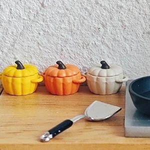 May include: Three miniature pumpkin-shaped containers in yellow, orange, and cream colours, each with a black stem and small handles. A small spatula and a black pan are also visible, suggesting a kitchen or food-related theme.