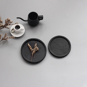May include: Two black wooden coasters with a small sprig of dried flowers on one of the coasters.