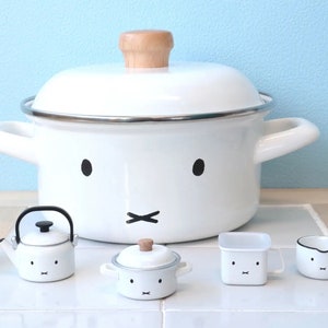 May include: A set of white enamel cookware with black and brown details. The set includes a large pot with a lid, a smaller pot with a lid, a kettle, a small saucepan, and a square container. All of the pieces have a cute cartoon face design.