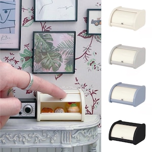 Könnte beinhalten: Vier Miniatur-Brotkästen in verschiedenen Farben: Weiß, Hellgrau, Hellblau und Schwarz. Die Brotkästen stehen auf einem weißen Regal mit einem weißen Tisch und einem kleinen Radio auf der linken Seite. Die Brotkästen haben eine abgerundete Oberseite und einen kleinen Knopf an der Vorderseite.