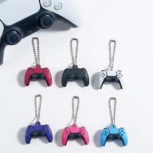 May include: Six miniature video game controller keychains with silver ball chains, arranged in two rows. The controllers come in red, black, white, purple, pink, and light blue, each featuring black joysticks and buttons, positioned below a full-sized controller.