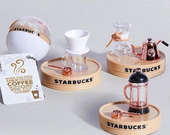 Może przedstawiać: Zestaw pięciu miniaturowych figurek Starbucks z motywem kawy. Zestaw zawiera kulkę z ziarnami kawy, przelewowy ekspres do kawy, prasę francuską, dzbanek do kawy i karafkę. Każda figurka stoi na małej, okrągłej podstawie z logo Starbucks.