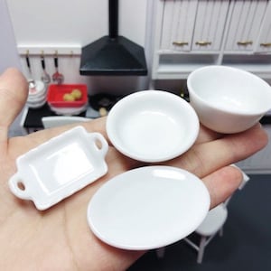 May include: A set of four white ceramic miniature dishes, including a small bowl, a larger bowl, a rectangular dish with handles, and a round plate. The dishes are arranged on a hand.