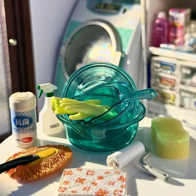 May include: A miniature cleaning set featuring a teal bowl with a spoon, yellow gloves, a spray bottle, a roll of lint remover, a sponge, a cleaning brush, and a container with Japanese text. The set is arranged on a white surface.