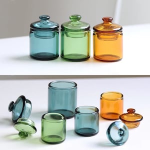 May include: Set of six glass jars with lids in various colours, including teal, green, and amber. The jars are of different sizes and are arranged on a white surface.