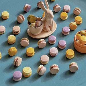 以下が含まれることがあります： 青い表面に、パステルカラーのマカロンが置かれています。セラミック製のウサギの置物が、マカロンの巣を持っています。マカロンは、ピンク、イエロー、パープルなど、さまざまな色合いです。