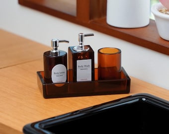 Könnte beinhalten: Ein braunes Badezimmer-Set aus Glas mit silbernen Pumpenspendern und einem kleinen Becher. Das Set enthält einen Spender für Körperlotion und Duschgel und wird auf einem passenden Tablett präsentiert. Die Etiketten lauten "Body lotion" und "Body Wash".