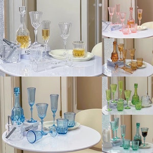 May include: A set of six clear glass decanters and glasses in various colours, including blue, green, pink, and orange. The decanters and glasses are arranged on a white table with a Kinfolk magazine in the background.