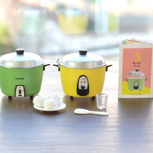 May include: Two miniature rice cookers, one green and one yellow, with a small plate of white rice balls, a measuring cup, a rice paddle, and a small cookbook with the text "3 Recipe Book" on the cover.