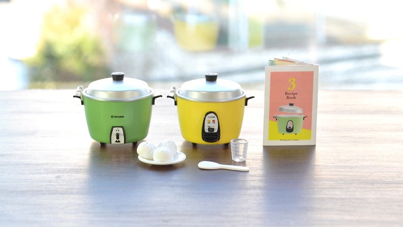 May include: Two miniature rice cookers, one green and one yellow, with a small plate of white rice balls, a measuring cup, a rice paddle, and a small cookbook with the text "3 Recipe Book" on the cover.