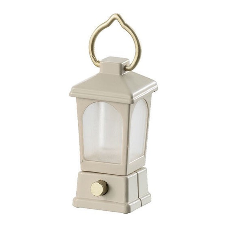May include: A small, beige lantern with a gold handle and a clear glass window. The lantern has a small, round knob on the bottom.