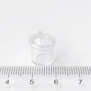 May include: A miniature, transparent glass jar with a lid. The jar is cylindrical with a rounded lid and a small knob on top. A ruler is in the foreground, with measurements in centimetres.
