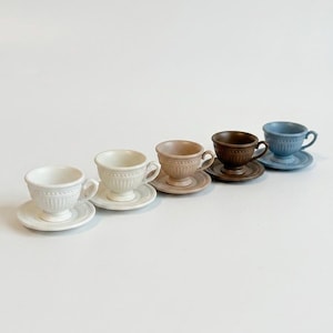 May include: Five ceramic teacups and saucers in a row. The cups and saucers are in shades of white, beige, brown, and blue. Each cup has a handle and sits on a matching saucer. The cups have a ribbed design.