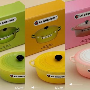 Puede incluir: Tres cocottes de hierro fundido Le Creuset en verde, amarillo y rosa, cada una con un pomo negro y asas laterales. Las cajas y cocottes están etiquetadas con el logotipo de Le Creuset y el texto "MADE IN FRANCE". Cada cocotte mide 6,5 cm de diámetro.