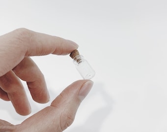 May include: A small, clear glass bottle with a cork stopper. The bottle is being held by a hand.