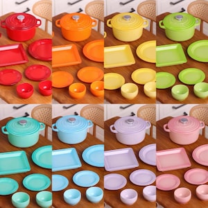 Puede incluir: Un juego de diez utensilios de cocina y vajilla en miniatura de varios colores, incluyendo rojo, naranja, amarillo, verde, azul, morado y rosa. El juego incluye una olla pequeña con tapa, un plato rectangular, un plato redondo, un tazón pequeño y una taza pequeña. El juego está dispuesto sobre una superficie de madera.